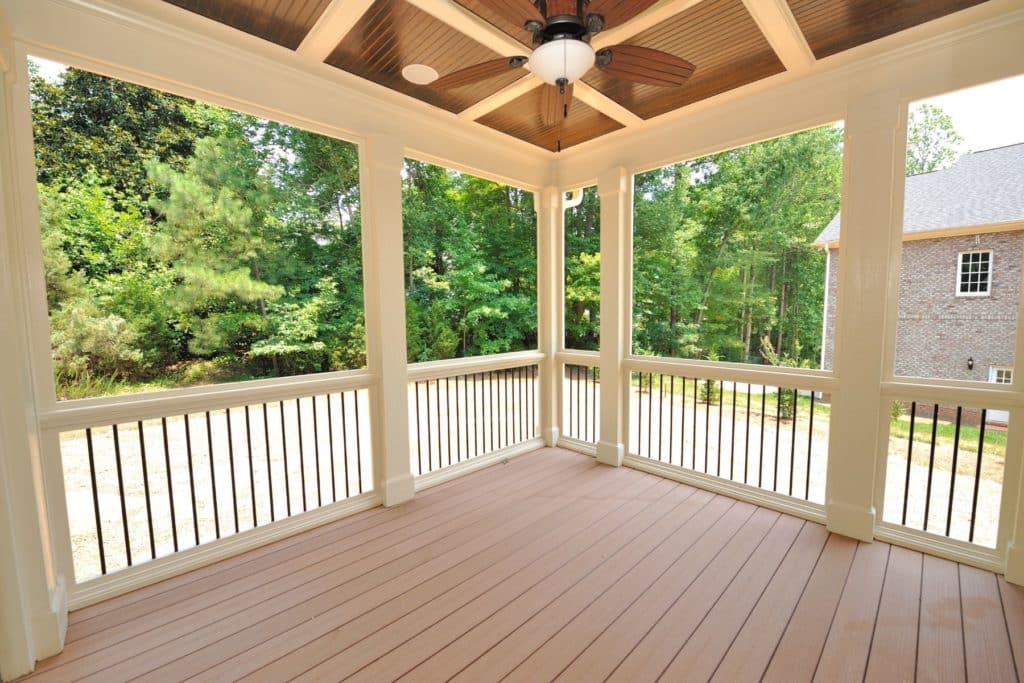 Spacious covered porch with a wooden floor, white columns, and railings, overlooking green trees, with a ceiling fan above.